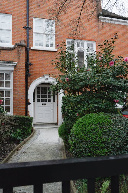 A row of lush, dense lavender bushes with light purple flowers lines the front of a red-brick residential building in a suburban garden setting. The bushes are well-maintained, with some trimmed to ke