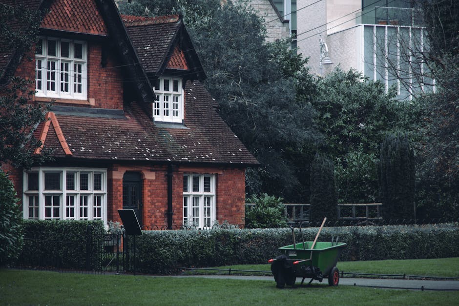 A semi-detached red brick house with a steep, tiled roof and white-framed, multi-pane windows is surrounded by a well-maintained garden featuring a lush, trimmed lawn and bordered by neatly shaped hed